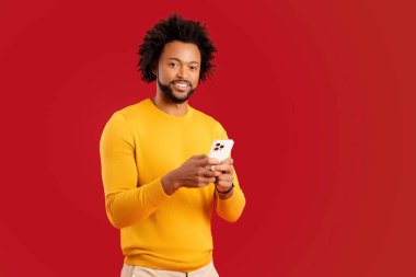 Mutlu genç adam akıllı telefon ve mesaj kullanıyor, sosyal ağlarda mesajlaşıyor, pozitif Afrikalı Amerikalı adam mobil uygulama kullanıyor, kırmızı arka planda izole edilmiş bir stüdyo resmi.