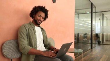 Yakışıklı, Afro-Amerikalı, serbest çalışan, yeşil tişörtlü, iş yerinde dizüstü bilgisayarda çalışan, uzaktan çalışmanın ve çalışma saatlerinin keyfini çıkaran, kameraya bakıp gülümseyen,