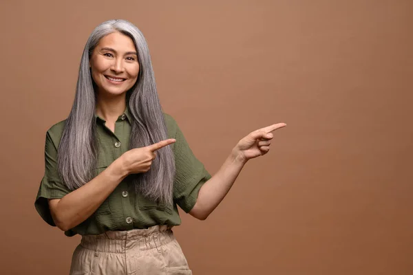 笑顔グレー髪40歳アジアの女性ポインティング離れて上の空白のコピースペース ショー場所のためにあなたの広告隔離された上茶色の背景 — ストック写真