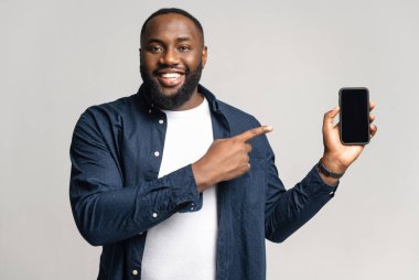 Elinde akıllı telefon olan heyecanlı Afrikalı adam gri arka planda izole edilmiş, telefonu boş ekran ile işaret ediyor, mobil uygulama reklamı yapıyor, anlaşmayı gösteriyor.