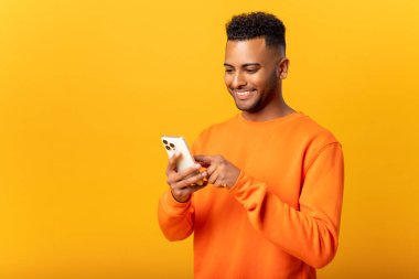 Ecstatic young Indian man standing isolated on yellow and using trendy smartphone, looking at screen and smiling joyfully, millennial guy enjoy new mobile application features