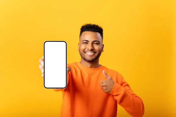 英俊年轻人的肖像展示了带有空白屏幕的智能手机 并展示了大拇指 男人喜欢移动应用 微笑着看着相机 感到满意 — 图库照片