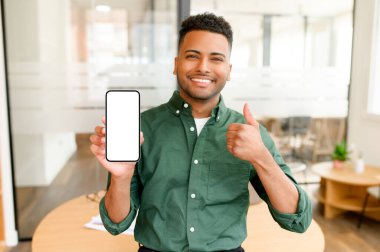 Yakışıklı genç Hintli adamın portresi akıllı telefonu boş ekranla gösteriyor ve baş parmağını kaldırıyor, mobil uygulamayı seviyor, serbest çalışan biri kameraya bakıp gülümsüyor, tatmin oluyor.