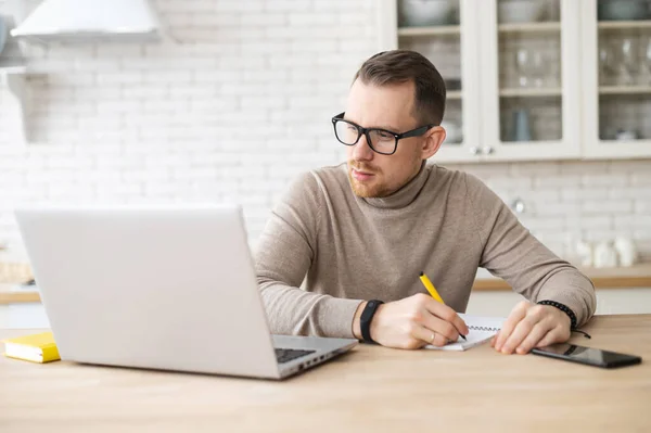 焦点を当てたハンサムな若い男は ペンで書く ノートパソコン上のオンラインウェビナー ビデオクラスを見て オンラインで勉強しています 自分の家に座って研究をしています — ストック写真