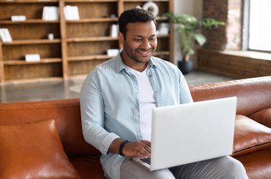 Keyifli, mutlu Hintli adam dizüstü bilgisayarı kullanarak arka planda kitap raflarıyla oturuyor koyu renk saçlı İspanyol internette sörf yapıyor, boş zamanlarını sosyal ağda geçiriyor.