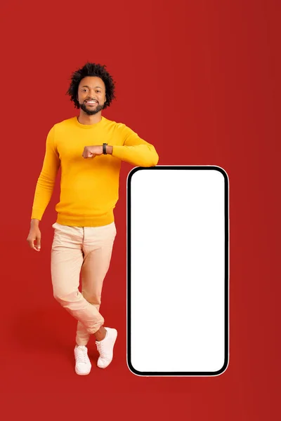 快乐而自信的非洲裔美国年轻人靠在巨大的智能手机上 屏幕空空如也 背景是红色的 卷曲的小伙子脸上挂着灿烂的笑容 还有手机应用程序的广告 全身上下都是 — 图库照片