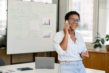 Son derece yetenekli bir iş kadını, koç, gözlüklü bir yönetici arka planda beyaz tahtayla birlikte çalışarak akıllı telefonla konuşuyor, motive edici bir sunum hazırlıyor, çalışanlar için iş planı yapıyor.