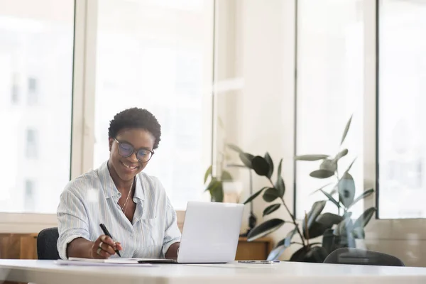 Inspired smiling female student, freelancer, office employee in glasses using the laptop for remote work, making research and taking notes, african-american business woman in modern coworking space