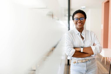 Kararlı ve gururlu bayan girişimci ofis koridorunda kollarını kavuşturarak duruyor, modern ve stil sahibi, kısa saçlı, iş yerinde kameraya bakan serbest bir kadın.