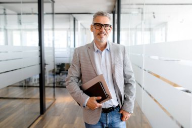 Ofis koridorunda dizüstü bilgisayar taşıyan orta yaşlı, karizmatik bir adamın portresi. Yakışıklı, olgun sakallı iş adamı, dişlek gülümsemesiyle kameraya bakıyor.