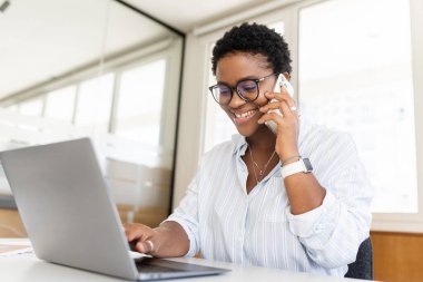 Laptop kullanan kendine güvenen genç girişimci, çağdaş çalışma alanında cep telefonuyla konuşuyor. Kısa saçlı, şık bir Afro-Amerikan kadın masada oturuyor ve hoş bir telefon konuşması yapıyor.