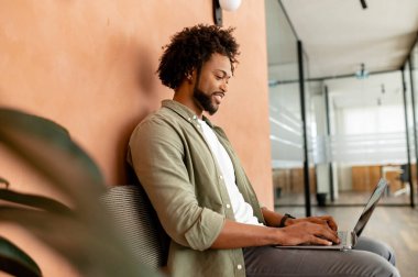 Afro-Amerikalı erkek çalışan, serbest çalışan, ofis koridorunda dizüstü bilgisayarda yazan programcı, olumlu ve gülümseyen takım üyesi e-postalara cevap veriyor, iş arkadaşlarıyla sohbet ediyor.