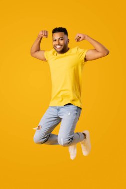 Neşeli, atletik Hintli adamın uzun boylu portresi yükseğe zıplayan ve pazılarını gösteren, sarı arka planda izole edilmiş genç adam zafer işaretinde yumruklarını kaldırıyor.