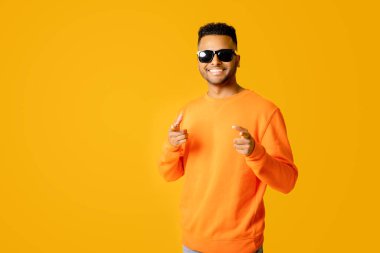 Havalı, yakışıklı, turuncu kazaklı ve güneş gözlüklü Hintli genç adam, güneşli bir günün keyfini çıkarıyor, sarı arka planda izole bir şekilde duruyor, kamerayı işaret ediyor.