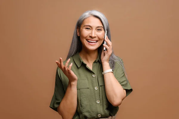 明るく幸せな幸せな成熟したアジアの女性は 茶色の背景で隔離されたスマートフォンで話します 楽しい電話会話を持っている40代女性 友人とチャット — ストック写真