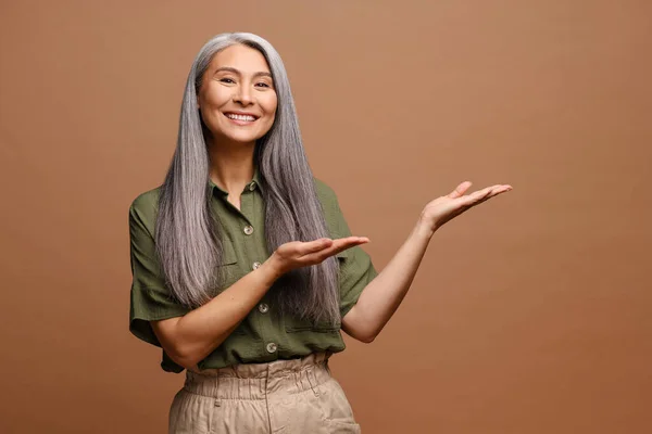 コピースペースで手を指す灰色の髪のアジア女性の肖像画 広告のための領域を示しています 茶色の背景に隔離された屋内スタジオショット — ストック写真
