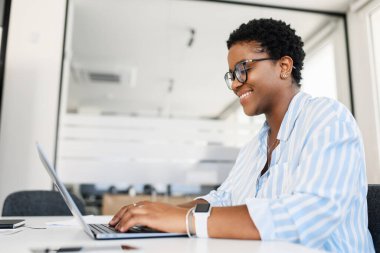 Modern çalışma ortamında dizüstü bilgisayar kullanan iyimser Afro-Amerikan kadın ofis çalışanı, bilgisayar üzerinde çalışan, ekranda seyreden ve gülümseyen, yan gören iş kadını.