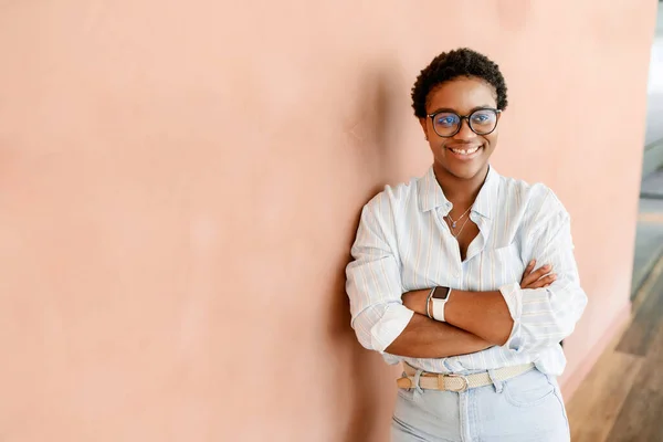 Accomplished african female entrepreneur confidently stands with arms folded, gazing at the camera with a smile. Optimistic and cheerful freelancer woman in a casual shirt working in shared workspace