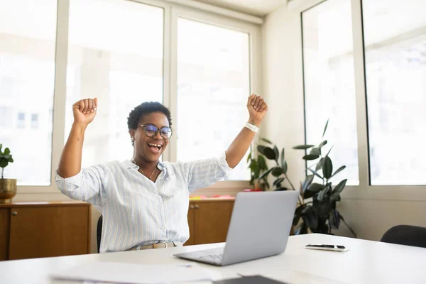 Excited African-American female freelancer raising hands in ecstatic, business woman got promotion, job offer, celebrating success, looking at laptop screen, screaming yes, triumph and victory concept