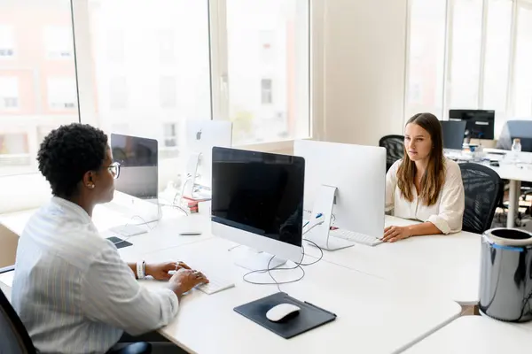 現代のオープンスペースオフィスで机に座っている2人の女性同僚 コンピュータで働くスマートで集中的な女性 職場のオフィス用品 フレンドリーな作業環境を楽しむ — ストック写真