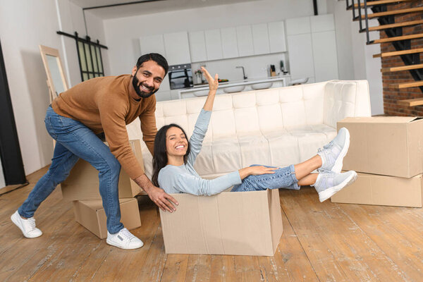Happy Indian couple having fun while unpacking belongings on moving day. Excited wife riding in cardboard box while her husband push it in new house apartment