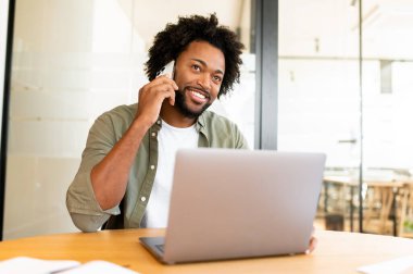 Laptop kullanan ve çağdaş çalışma alanında cep telefonuyla konuşan kendine güvenen genç girişimci. Kıvırcık Afro-Amerikan adam masada oturuyor ve hoş bir telefon konuşması yapıyor.
