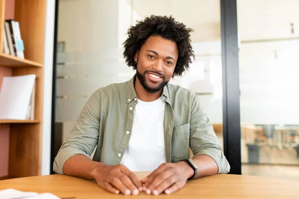 Portrait of handsome curly african-american sitting at the office desk and in front of the camera, looks at you with friendly smile, businessman involved in video conference, video call participant