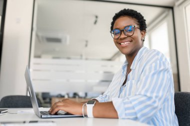 Ofisteki masada dizüstü bilgisayar kullanan Afro-Amerikalı bir iş kadını, gülümseyen karizmatik bir kadın çalışan, serbest çalışan, günlük mavi gömlekli ve gözlüklü biri.