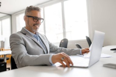Ofiste dizüstü bilgisayar kullanan kendine güvenen bir iş adamı. Dost canlısı 50 'lerin erkek çalışanı iş yerinde oturuyor, ekrana bakıyor, araştırıyor, analiz ediyor, yan görüş yapıyor.