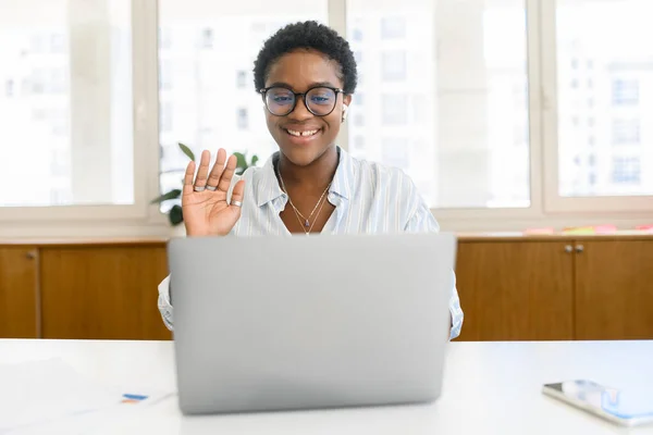 Cheerful and happy African-American woman wearing eyeglasses and shirt using a laptop for video meeting, video call, holding an online conference in the home office, female colleague greeting