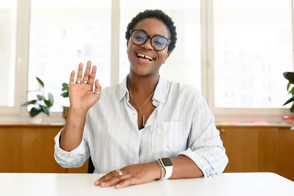 Young African-American female applicant involved online interview or video call, online teacher sitting at the desk and looking and waving at the camera, saying hello. Job hunting, e-learning concept