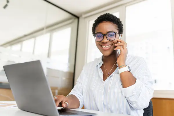 African-American female office employee in smart casual talking on the phone, and using a laptop on the workplace in modern office, female entrepreneur in stylish eyeglasses looking at camera