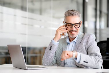 60 'ların kıdemli iyimser işadamı akıllı telefondan arıyor. Kol saatine bakıyor ya da modern ofisteki iş yerinde oturan akıllı bir saat. Dakiklik ve insanlar kavramı