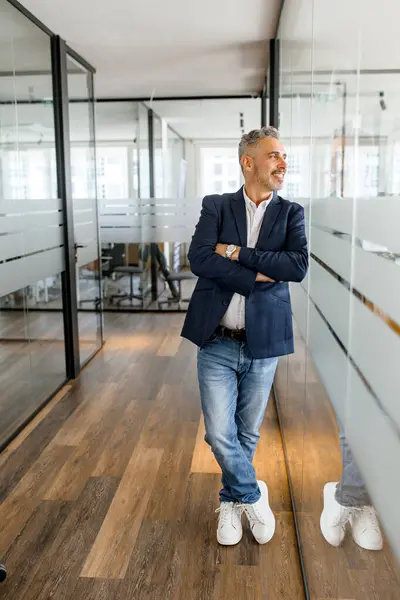 Ambitious mature male entrepreneur ceo manager stands in contemporary office space with arms crossed leaned on glass partition, smiling businessman looks aside, full length