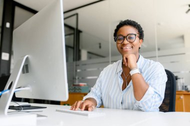 Pozitif Afro-Amerikan kadın çalışan, serbest çalışan ya da programcı bilgisayar başında oturmuş, yazı yazıyor, gülümseyen ve kameraya bakan bir kadın, bu şirkette çalışmaktan keyif alan bir programcı.