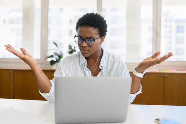 Puzzled African-American female office worker sitting in the workplace and shrugging shoulders, having problems with project, computer error, businesswoman in formal wear looking at computer monitor