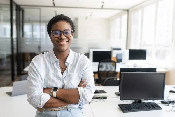 African-American female office employee with short hair standing with arms crossed in contemporary co-working office space, multiracial woman looking at camera. Diverse work team concept