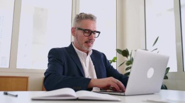 Kıdemli kıdemli gri saçlı işadamı ofis işi için dizüstü bilgisayar kullanıyor, olgun erkek çalışan e-postaları kontrol ediyor, müşterilerle iş yazışmaları yapıyor, kapalı alanda oturan kameralara bakıyor.