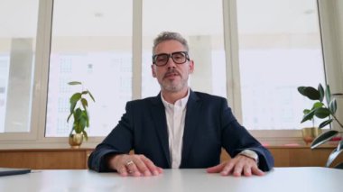 İyimser, olgun iş adamının portresi kameraya bakıyor ve gülümsüyor, el sallıyor, erkek çalışan müdürü sanal toplantının katılımcılarını selamlıyor. Video çağrısı
