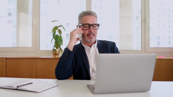 フレンドリー笑顔成熟したビジネスマンの男性従業員は 現代のオフィススペースに座って楽しい電話会話を持っています スマートフォンで話す陽気な灰色の髪のボス — ストック動画