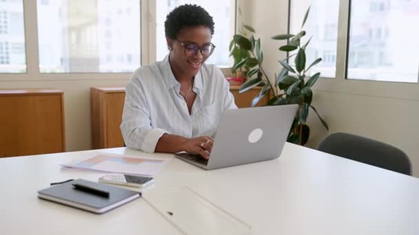 非洲裔美国女商人坐在办公室的办公桌前 微笑迷人的女雇员 身穿休闲蓝衬衫和眼镜工作的自由职业者的侧面形象 — 图库视频影像