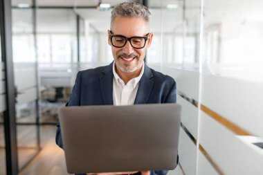 Zeki ve zeki 60 'lı yılların kıdemli iş adamı resmi takım elbise ve beyaz gömlek giyiyor. Kapalı alanda dizüstü bilgisayar kullanıyor. Erkek ofis çalışanı elinde bilgisayar tutuyor ve mesaj yazıyor.