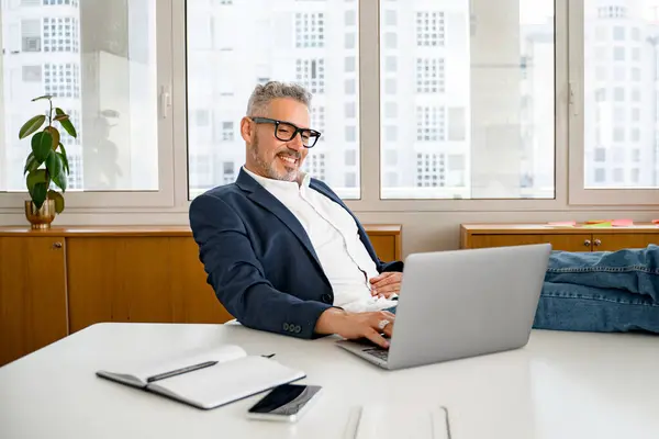 Self-assured 60s gray-haired modern confident businessman working in a comfortable and modern office in relaxed pose, putting his feet on the desk, successful mature business owner enjoying his job