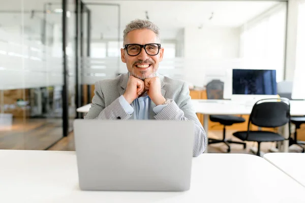 Optimistic mature male office employee sitting at the desk in front of laptop PC, looking at camera and smiling, successful senior businessman in modern loft office, manager ceo on the workplace