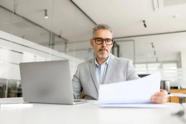 stock image An attentive businessman reviews documents while working on his laptop in a well-lit office space, portraying the meticulous nature of business planning and analysis