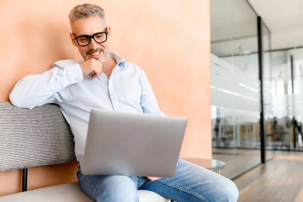 A mature businessman sits comfortably with a laptop on the couch in a casual work setting, reflecting the modern trend of flexible work environments, a balance between productivity and comfort concept