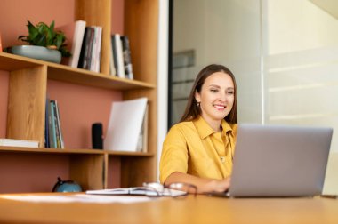 Konsantre gülümseyen kız modern ofiste çalışmak için dizüstü bilgisayar kullanıyor. Güzel genç bir kadın klavyeye yazı yazar ve bilgisayar ekranına bakar.