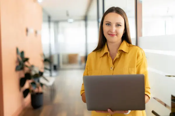 ラップトップを持つ笑顔の女性は カメラを見て 現代のオフィスで屋内で立っている カジュアルなウェアで陽気なビジネスマンは廊下に立っている電子メールをチェック — ストック写真