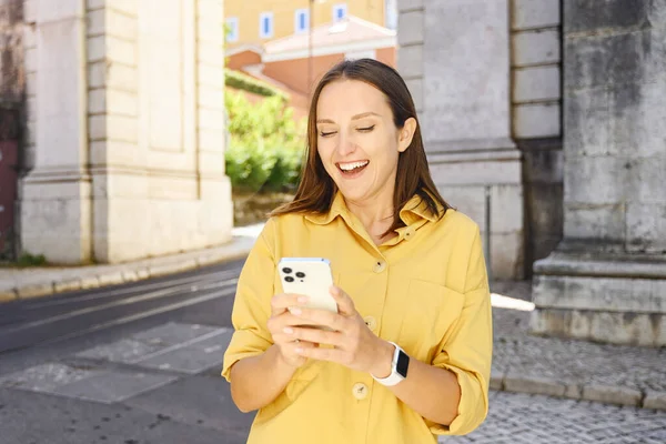 A delighted woman in a mustard yellow shirt and sunglasses laughs as she looks at her smartphone, with an arch and cobblestone street behind her, Lisbon, Portugal