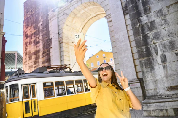 A joyful woman in yellow blouse and sunglasses poses with a peace sign while taking a selfie, with a historic arch and a classic yellow tram in the sunny background.